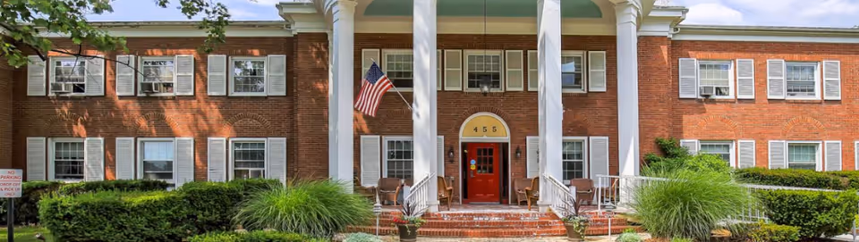 Front exterior view of a two-story brick building with white columns, white window shutters, and a red door. There is an American flag hanging near the entrance, surrounded by green shrubs and plants.