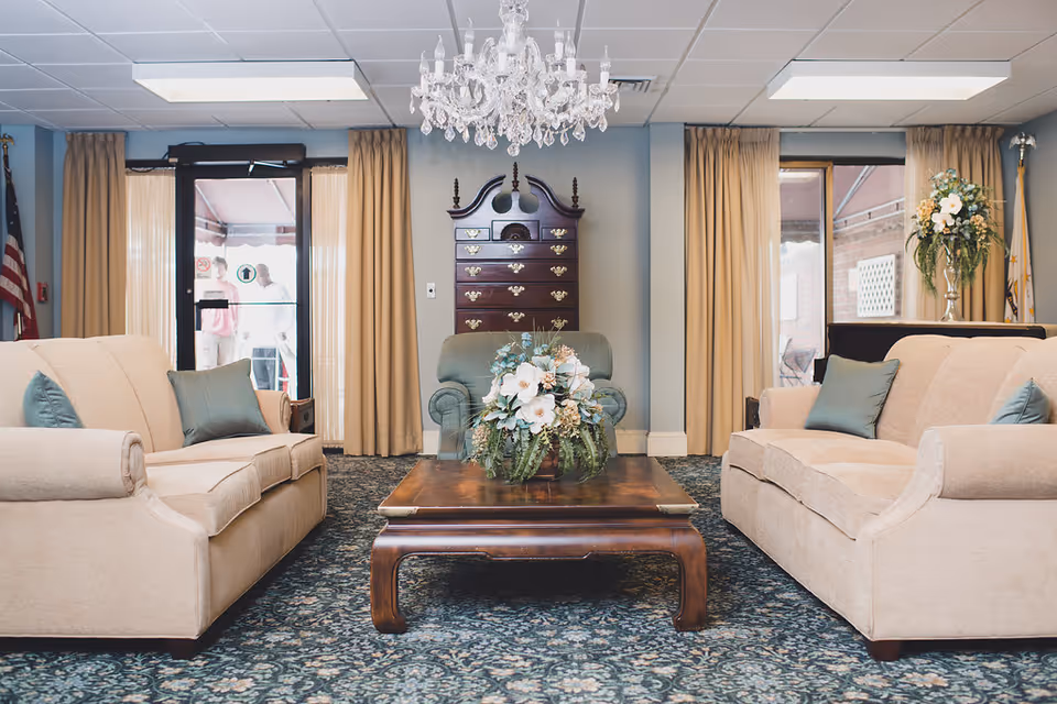 A cozy senior living center lounge area with two beige sofas facing each other, a wooden coffee table with a floral arrangement in the center, a green armchair against the wall, a tall wooden chest of drawers, beige curtains, and a crystal chandelier hanging from the ceiling.