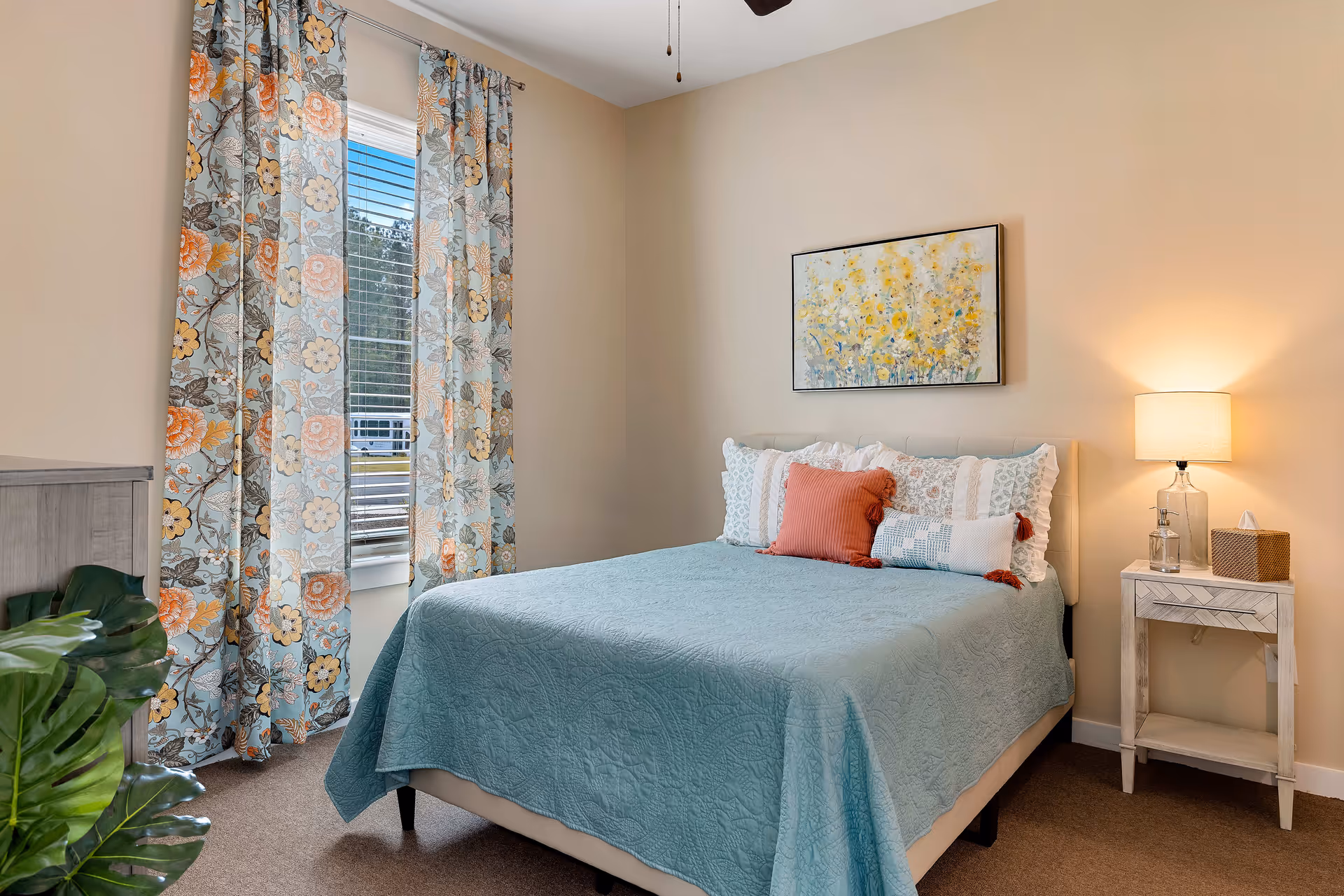 A tidy bedroom with a teal quilted bed, floral curtains at a window, a nightstand with a lamp, and framed wall art.