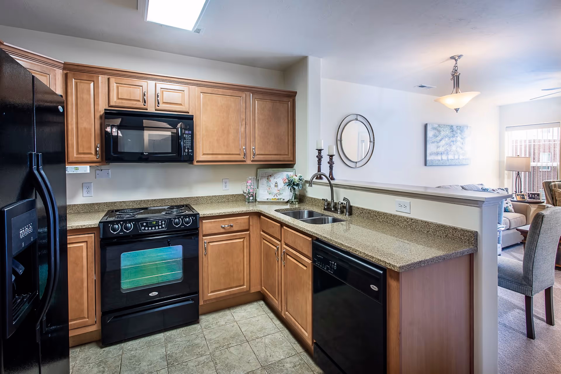 A modern kitchen with wooden cabinets, a black refrigerator, black microwave, black stove, and black dishwasher. The kitchen has a granite countertop with a double sink and a faucet. In the background, there is a dining area with a chair and a living room with a sofa, lamp, and wall art.