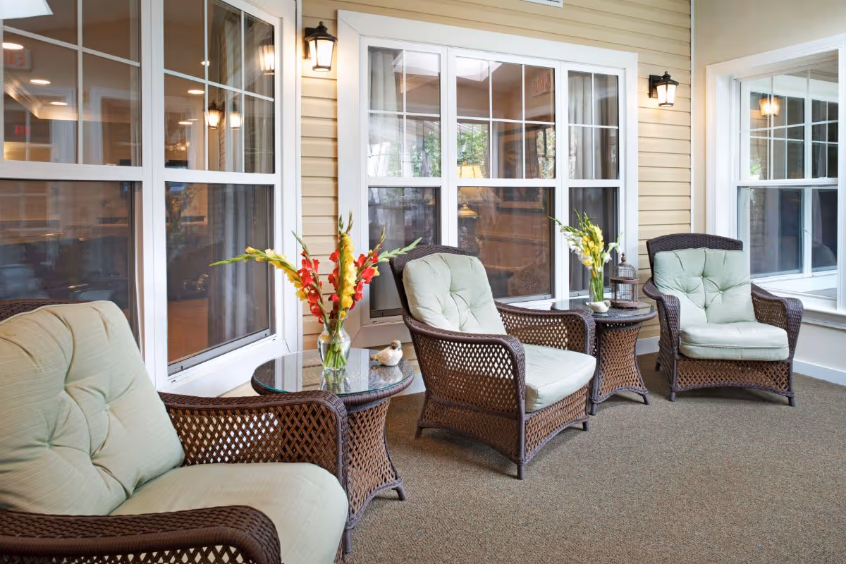 Sunlit indoor seating area with wicker armchairs, side tables, and vases of flowers in front of large windows.