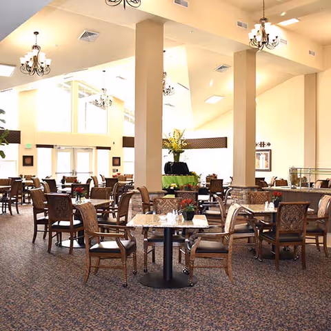 A spacious dining room with multiple tables and chairs arranged neatly. The room features high ceilings with chandeliers, large windows allowing natural light, and decorative plants on some tables. The carpet has a patterned design, and the walls are painted in a light color.