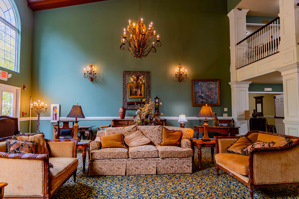 A spacious and elegantly decorated senior living facility common area with high ceilings, green walls, and a large chandelier. The room features a patterned carpet, a central floral arrangement on a wooden table, and multiple upholstered sofas and armchairs with cushions. There are wall sconces, table lamps, framed artwork, and a large window letting in natural light. A staircase and balcony are visible on the right side.