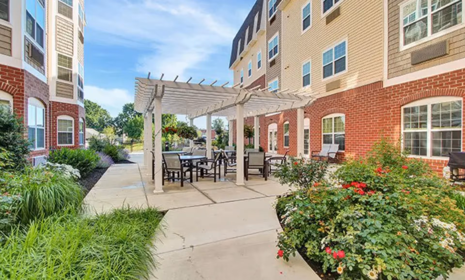 Outdoor courtyard with a white pergola and patio tables and chairs between brick-sided residential buildings, surrounded by landscaped plants and flowers.