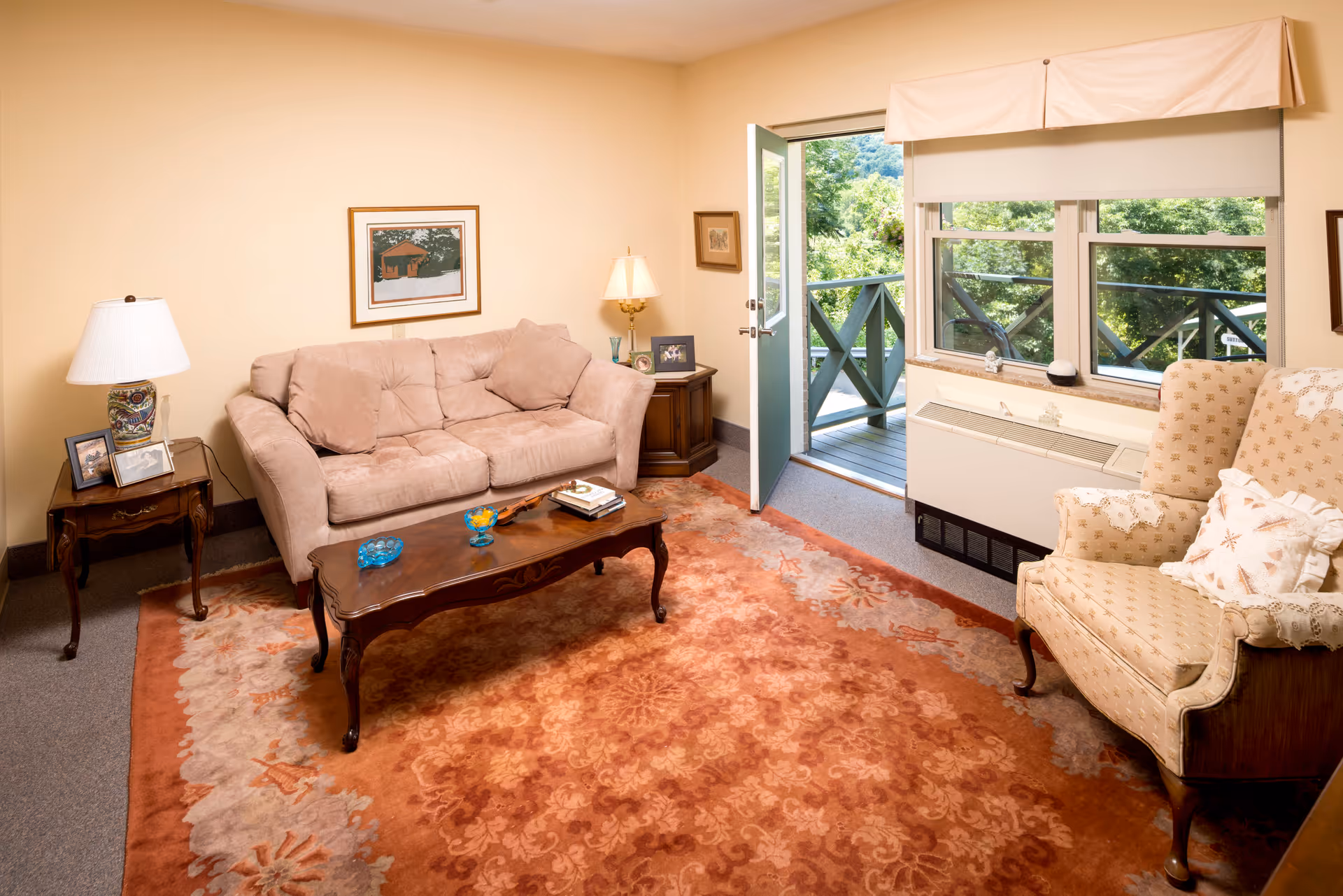 A cozy living room with a beige sofa, an upholstered armchair with pillows, a wooden coffee table with decorative items, and two side tables with lamps and framed photos. The room has a large patterned rug and an open door leading to a balcony with greenery outside.