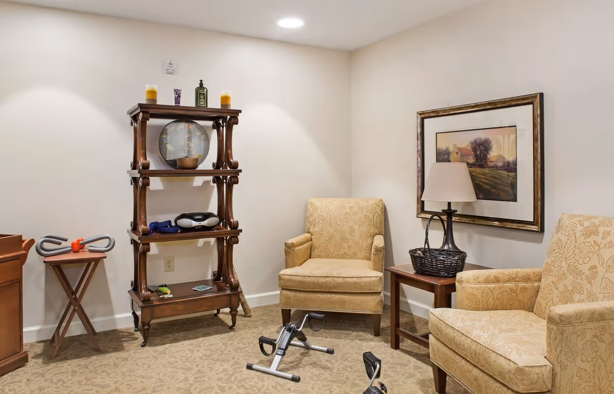 A cozy room with two beige patterned armchairs, a wooden side table with a lamp and a basket, a wooden shelving unit with decorative items, and a small exercise pedal machine on a carpeted floor. The walls are light-colored with a framed landscape painting.