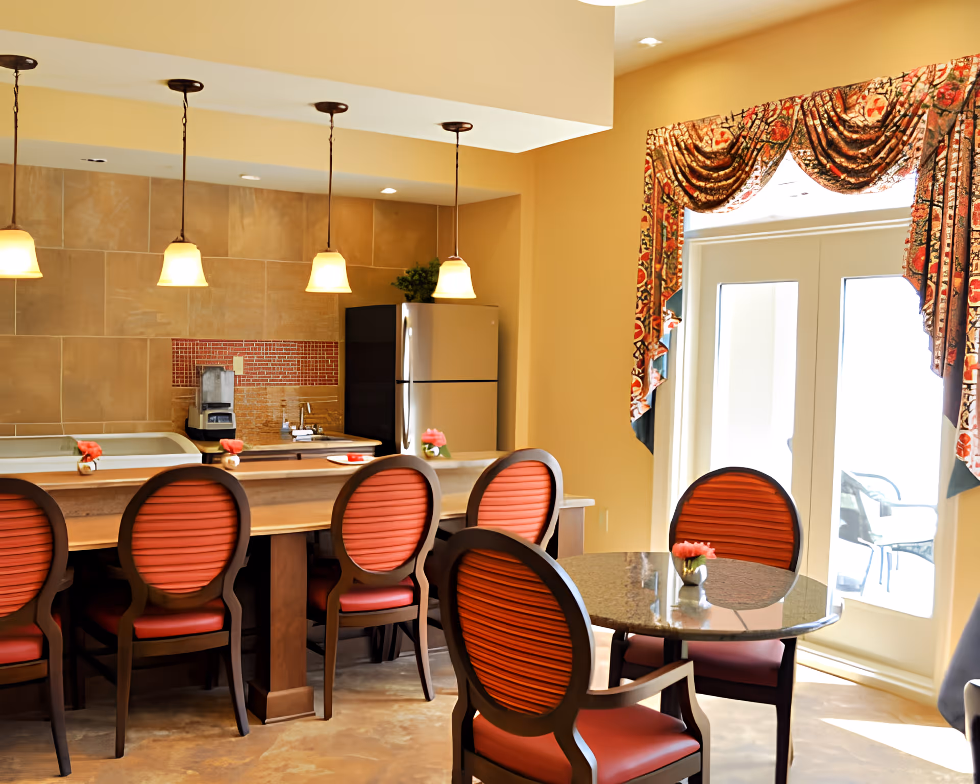 A bright communal dining and kitchen area with a long breakfast bar, red-upholstered chairs, pendant lights, and glass doors with patterned curtains.