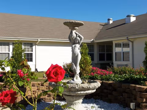 Statue fountain amid a rose garden beside a single-story senior living building exterior.