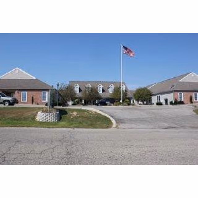 Exterior view of a single-story assisted living facility with a central driveway, an American flag on a flagpole, and a clear blue sky.
