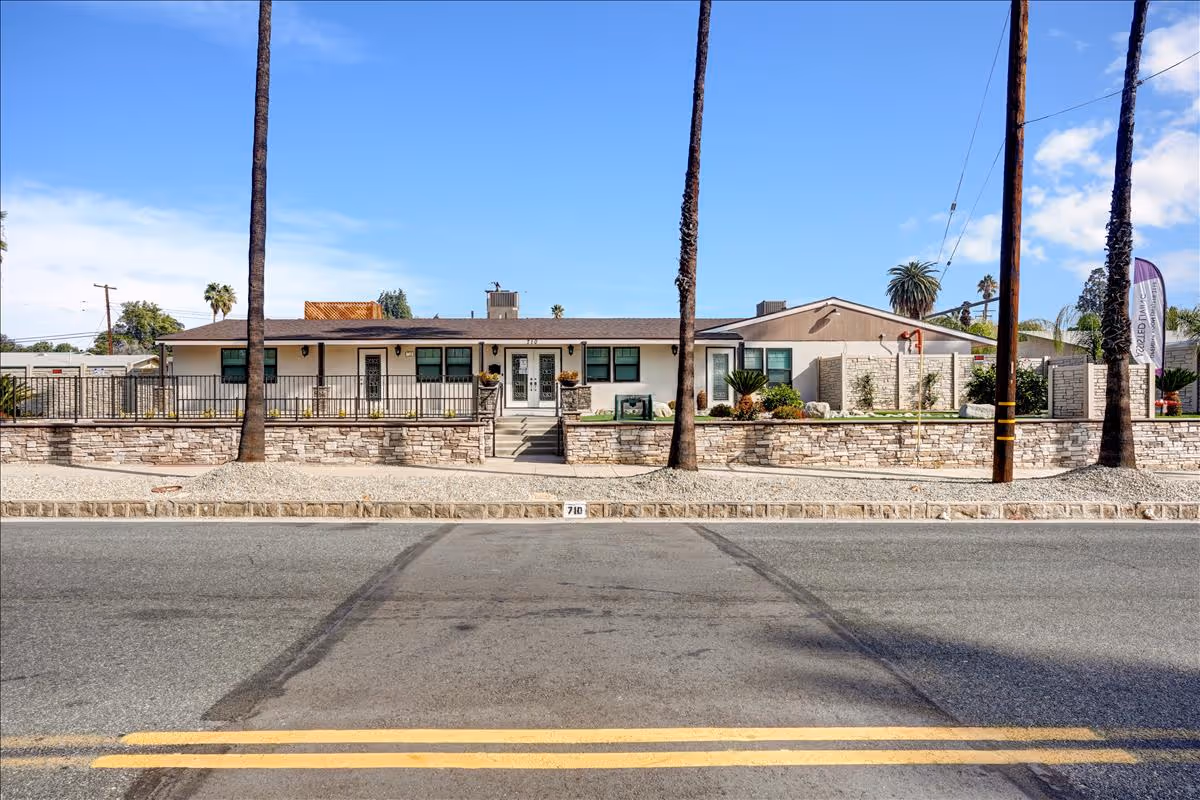 Front exterior view of a single-story building with a stone wall and steps leading up to the entrance. The building is surrounded by palm trees and has a clear blue sky in the background.