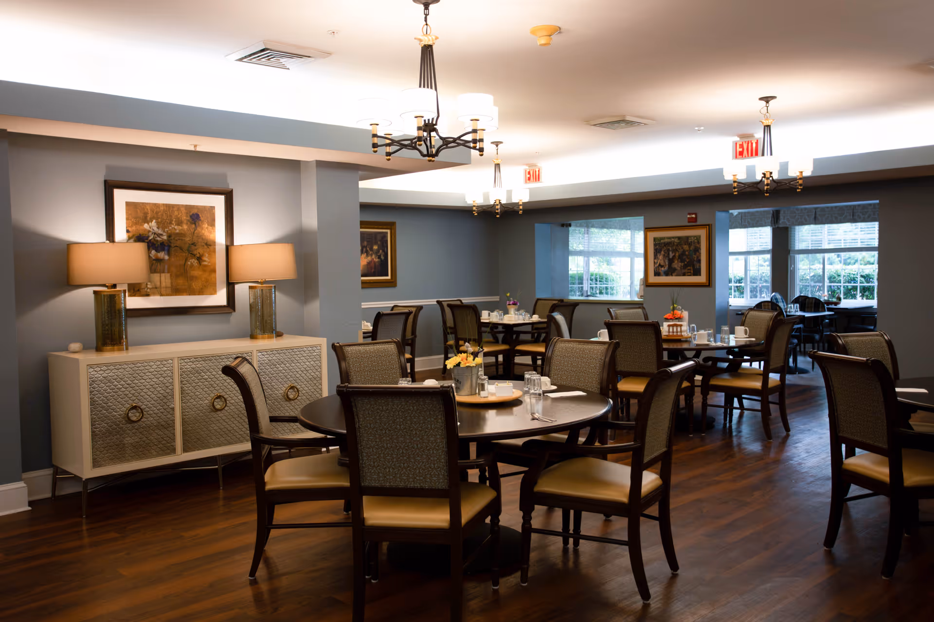 A well-lit dining room with several round tables and chairs arranged neatly. The room features wooden flooring, soft blue-gray walls, framed artwork, and two table lamps on a sideboard. Large windows allow natural light to enter, and ceiling light fixtures provide additional illumination.