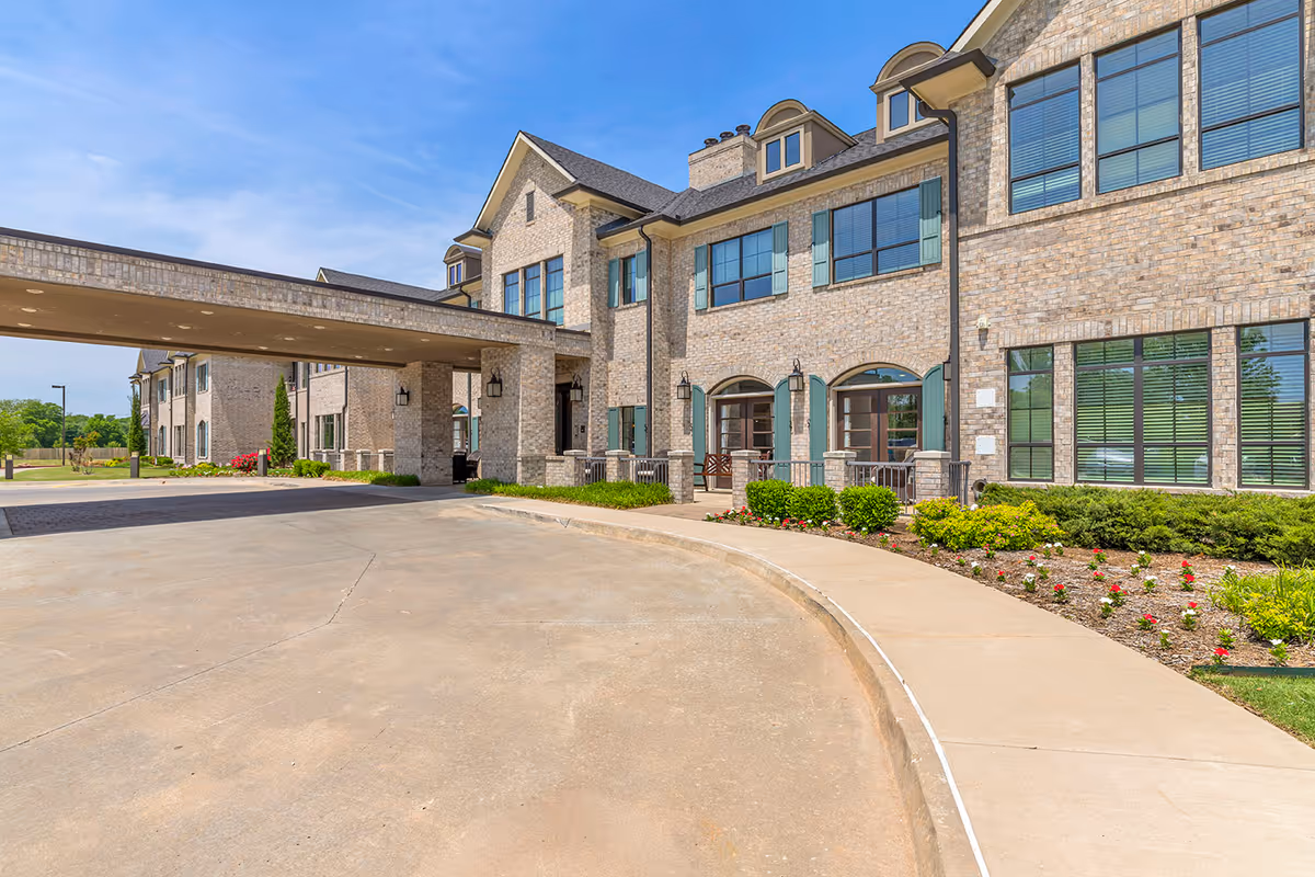 Exterior view of a senior living facility with beige brick walls, large windows with green shutters, and a covered driveway entrance. The building is surrounded by landscaped bushes and flowers under a clear blue sky.