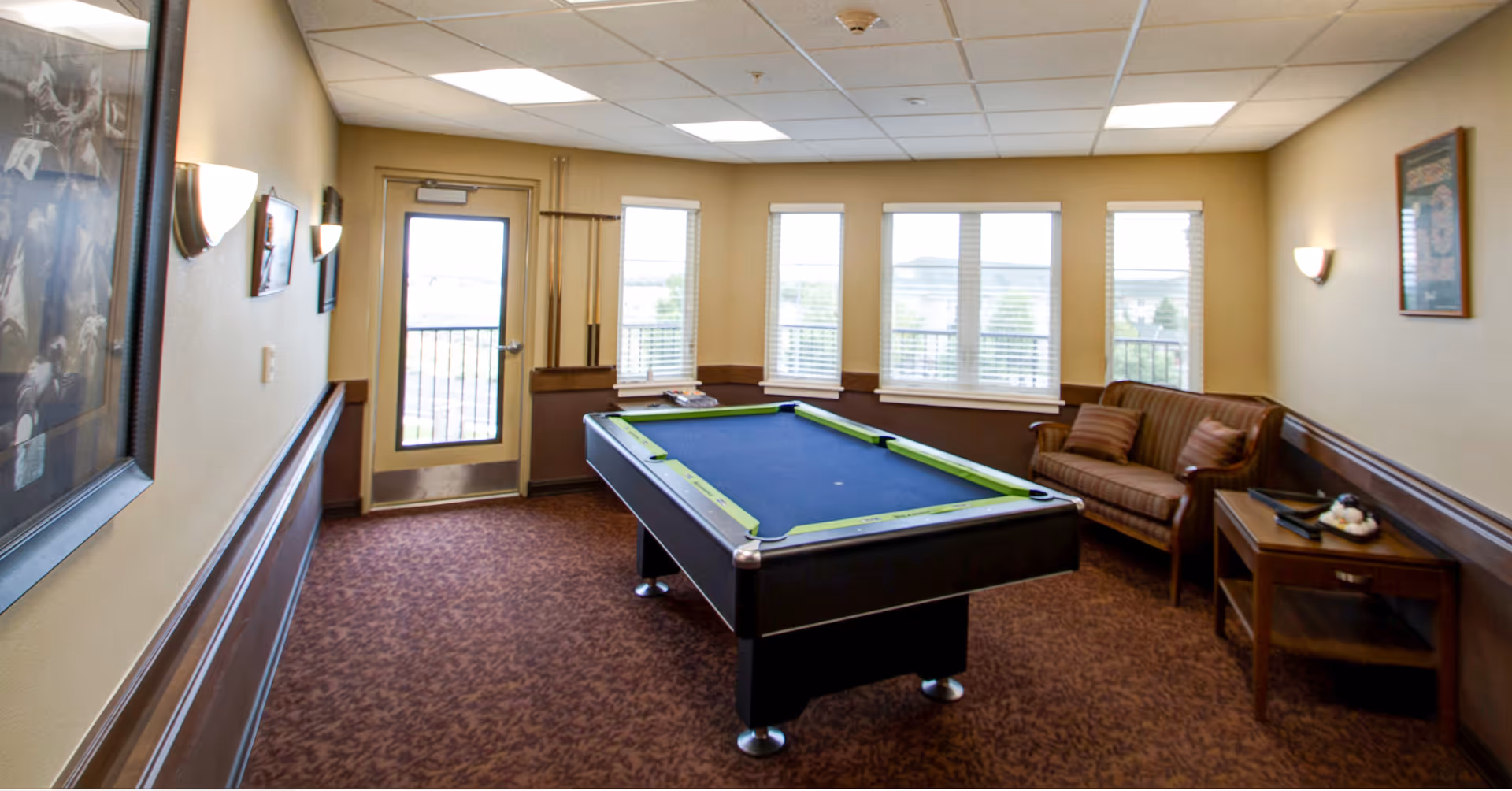 A recreational room with a pool table in the center, a brown patterned carpet, beige walls with wooden wainscoting, a brown couch with cushions, a side table with decorative items, several framed pictures on the walls, and windows letting in natural light. A door with a window leads outside.