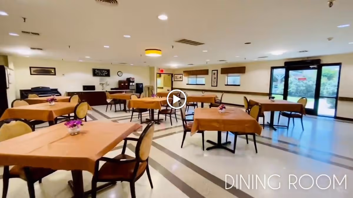 A spacious dining room with multiple tables covered with brown tablecloths, each table having a small flower arrangement. The room has beige walls, several windows with brown valances, and a glass door leading outside. The floor has a patterned design, and there are ceiling lights evenly spaced throughout the room.