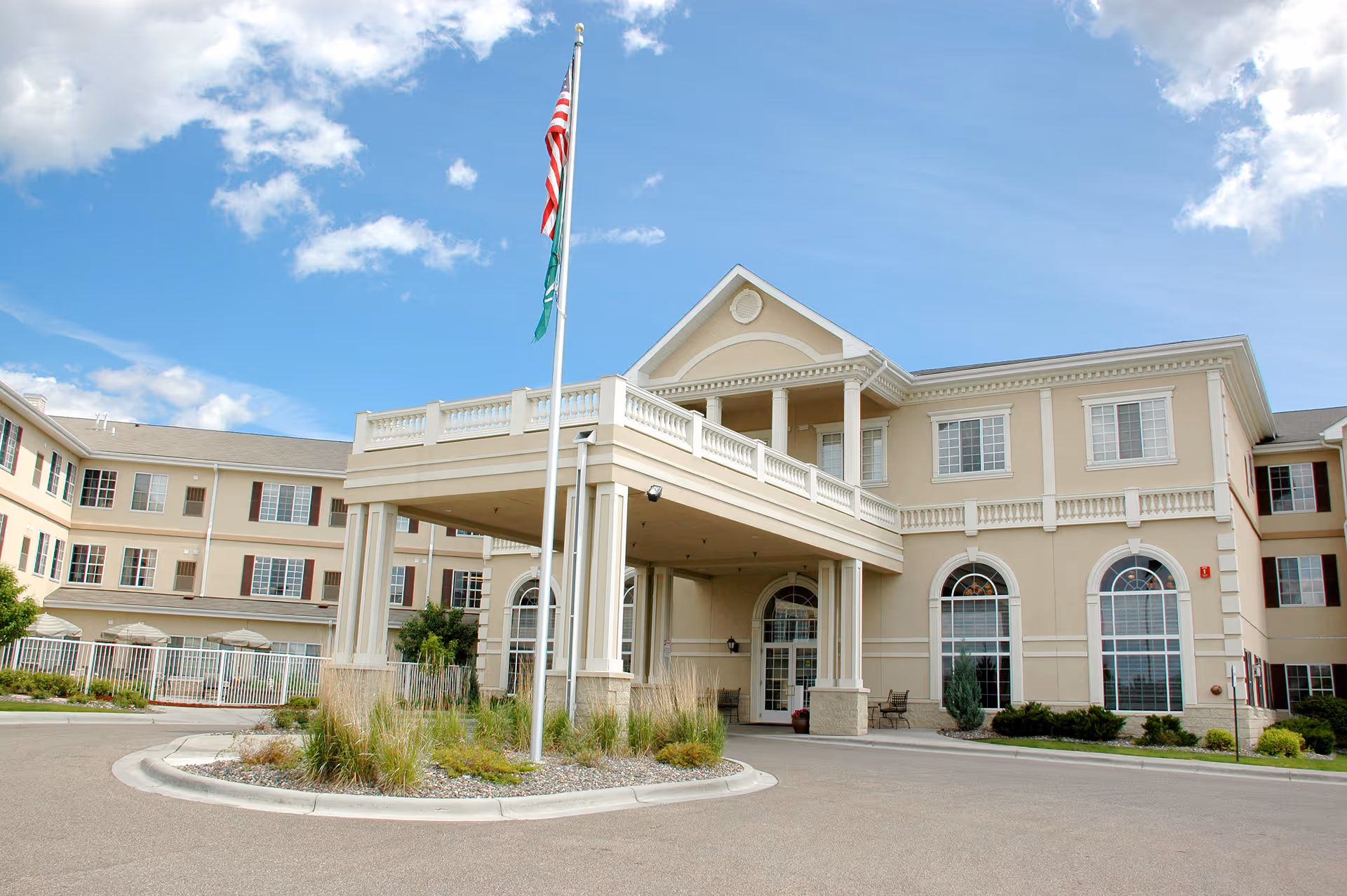 Exterior front view of a large, beige senior living facility building with multiple windows and a covered entrance supported by columns. There are two flagpoles with flags in front of the building, and landscaping with grass and shrubs around the entrance. The sky is partly cloudy with blue patches.