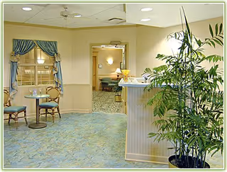 A bright senior living facility lounge with seating, a small table, a potted plant, and a reception-style counter leading to another sitting area.