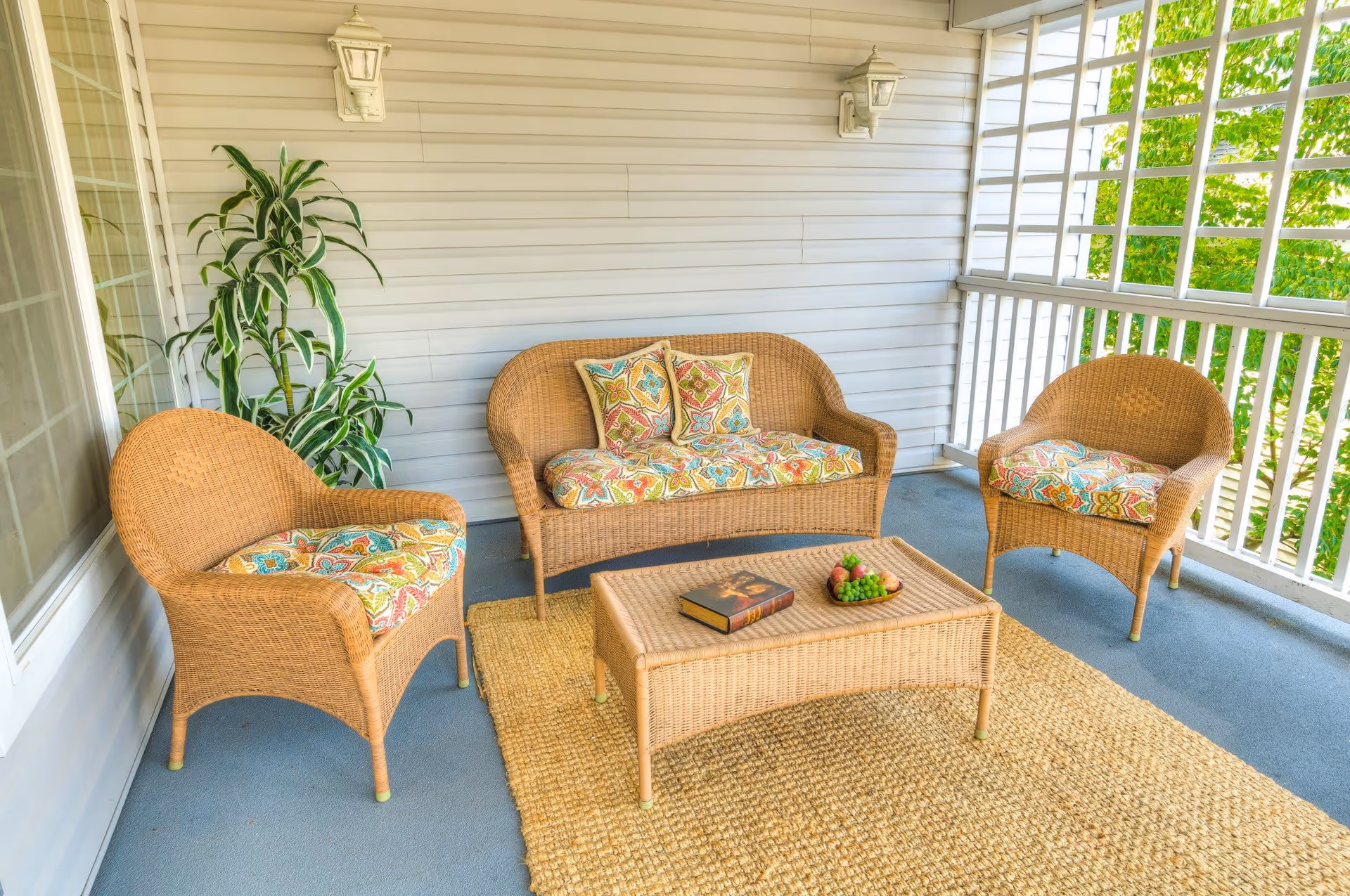 A cozy outdoor patio area with wicker furniture including a loveseat and two chairs, all with colorful patterned cushions. A wicker coffee table holds a book and a small plate of grapes. The patio is enclosed with white railings and has a beige woven rug on the floor. A green potted plant is placed against the wall, and two wall-mounted lantern-style lights are visible.