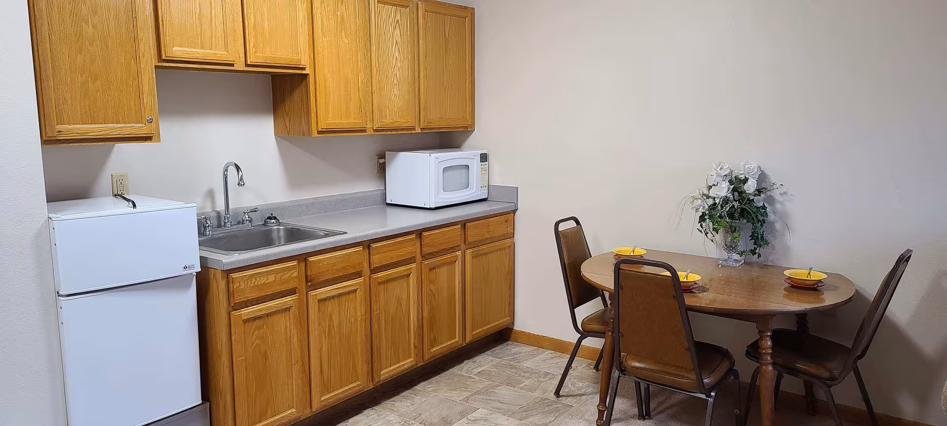 A small kitchen area with wooden cabinets, a countertop with a sink, a white microwave, and a white mini refrigerator. To the right, there is a round wooden dining table with three brown chairs and a vase with white flowers on the table. The walls are plain and light-colored.