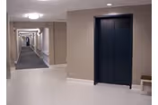 Interior hallway of a senior living facility with beige walls, a dark elevator door on the right, a bench beside the elevator, and a long corridor with handrails and carpeting extending into the distance.