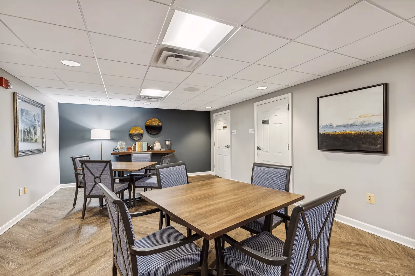 A well-lit interior room with wooden tables and cushioned chairs arranged for seating. The room has light-colored walls with framed artwork, a standing lamp, and a small shelf with decorative items and books against a dark accent wall. Two white doors are visible in the background.