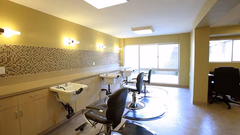 Interior view of a salon area in a senior living facility with three black salon chairs in front of white sinks mounted on a long beige countertop. The wall behind the sinks features a mosaic tile backsplash and is painted yellow. There are large windows letting in natural light and additional seating visible in an adjacent room.