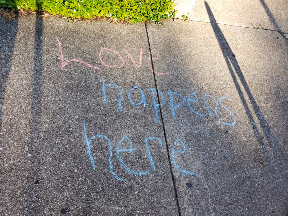 Concrete sidewalk with colorful chalk writing that reads "love happens here", shadows and greenery at the top edge.