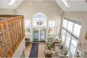 View from an upper level overlooking a spacious living room with high vaulted ceilings, large windows letting in natural light, a glass door entrance, comfortable seating including sofas and armchairs, a coffee table, and decorative plants.