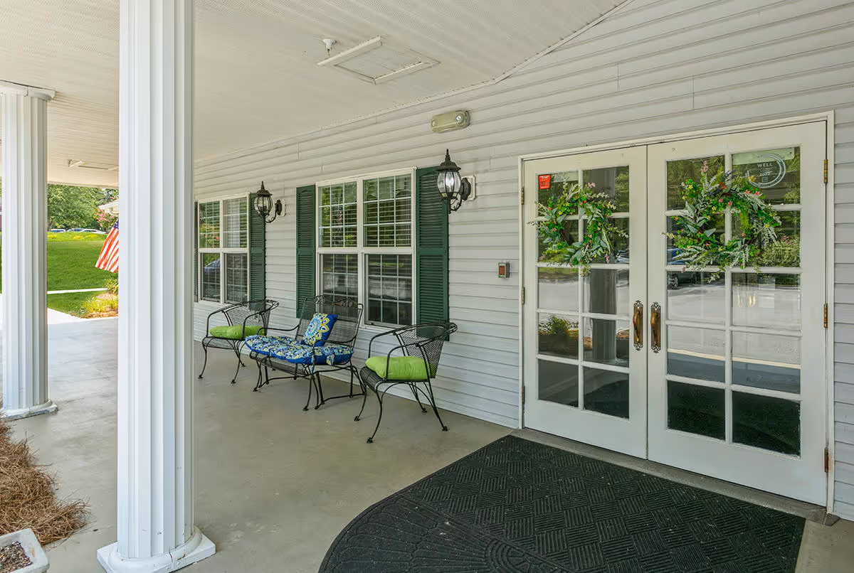 Covered porch area with white columns, black metal chairs with green and blue cushions, two wall-mounted lantern-style lights, and double glass doors decorated with green wreaths. An American flag is visible in the background on the left side.