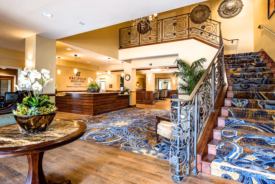 Bright and spacious senior living facility lobby with a wooden reception desk, decorative carpet, a staircase with ornate railing and patterned carpet, a round table with a floral arrangement, and warm lighting fixtures.