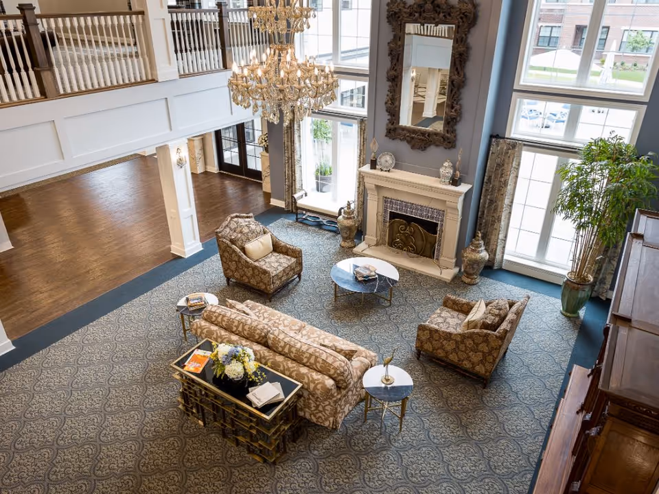 A spacious and elegant senior living facility common area with patterned carpet, a chandelier, a fireplace with a large ornate mirror above it, and comfortable upholstered seating including a sofa and two armchairs arranged around two small round tables. Large windows let in natural light, and there is a potted plant near the window.