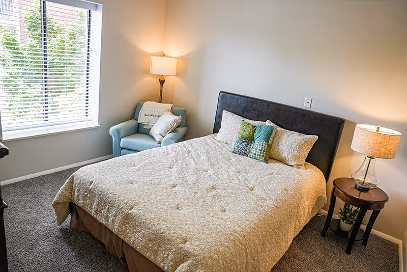 A cozy bedroom with a large bed covered in a beige patterned comforter and several decorative pillows. To the right of the bed is a wooden nightstand with a glass lamp and a small plant underneath. In the corner near the window, there is a light blue armchair with two pillows. The room has beige walls, a carpeted floor, and a window with white blinds letting in natural light.
