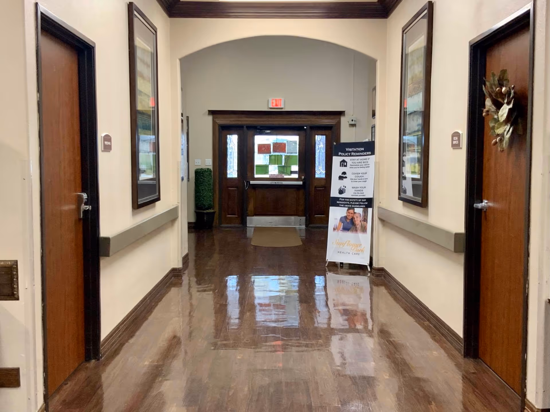 Polished wood-floor hallway with doors, framed artwork, and a visitation policy sign leading to double entry doors.