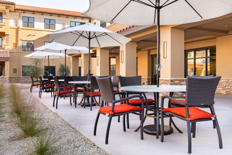 Outdoor patio area at Clarendale Of Chandler with multiple round tables, each surrounded by black wicker chairs with red cushions. Large white umbrellas provide shade over the tables. The patio is adjacent to a beige building with stone accents and large windows.