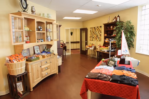 Bright interior of a senior living facility boutique showing display tables with folded clothes and snacks, a wooden hutch, and decorative items.