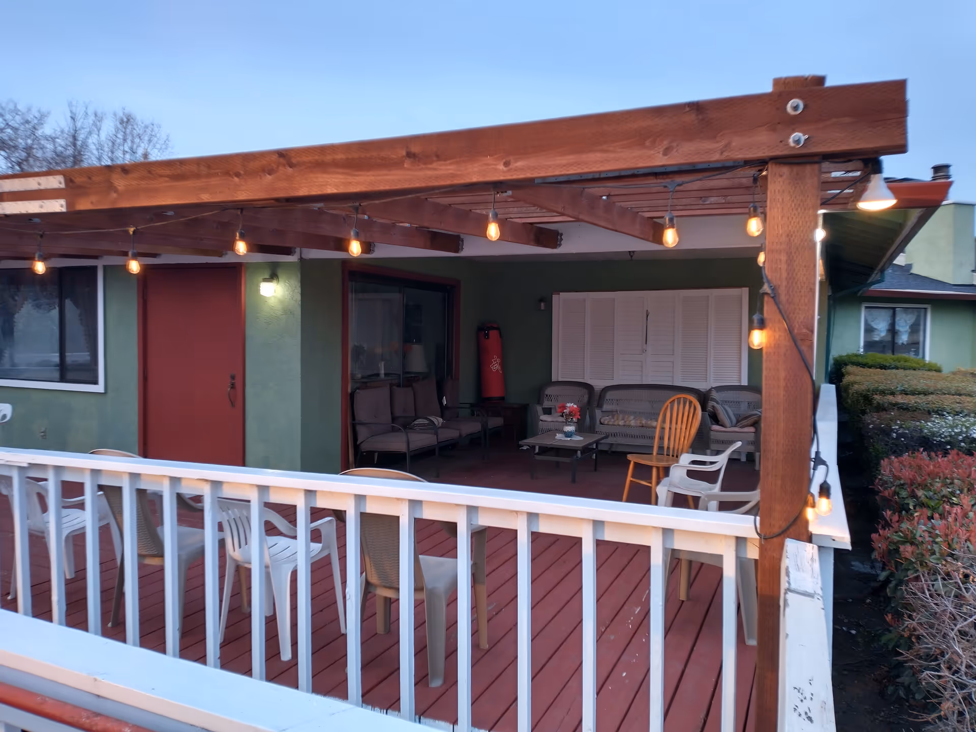 Outdoor patio area with a wooden pergola overhead adorned with string lights. The patio has a red wooden floor and white railing. There are several chairs, including plastic and wooden ones, arranged around a small table with a flower vase. The background shows a green building wall with a red door and windows.
