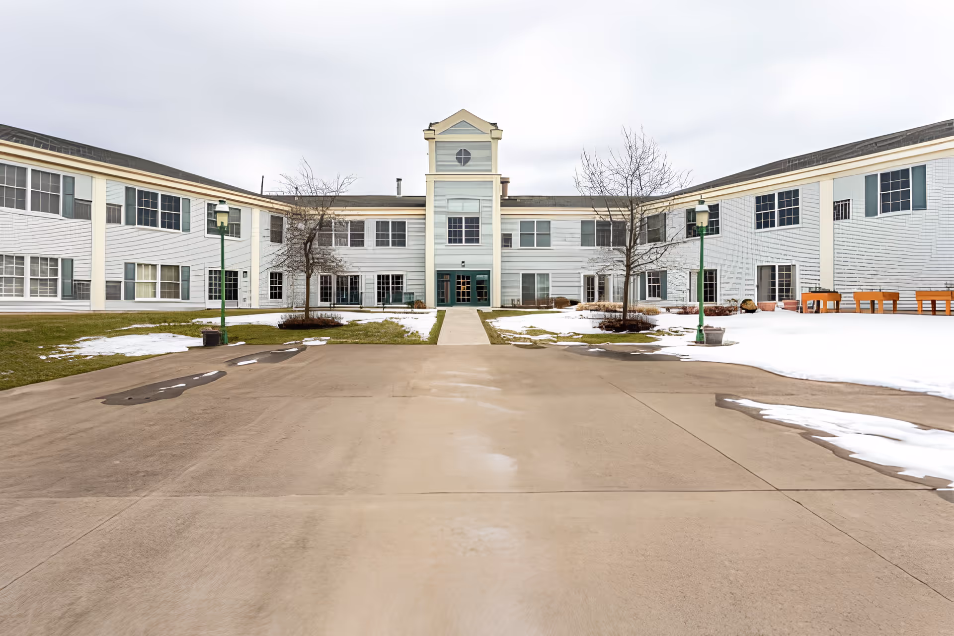 Front courtyard and main entrance of a two-story senior living building with patches of snow on the lawn.