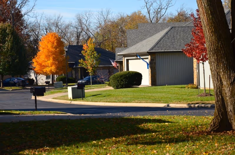 A suburban neighborhood street with houses, green lawns, and trees with autumn foliage in shades of orange, red, and green. The sky is clear and the scene is well-lit by sunlight.