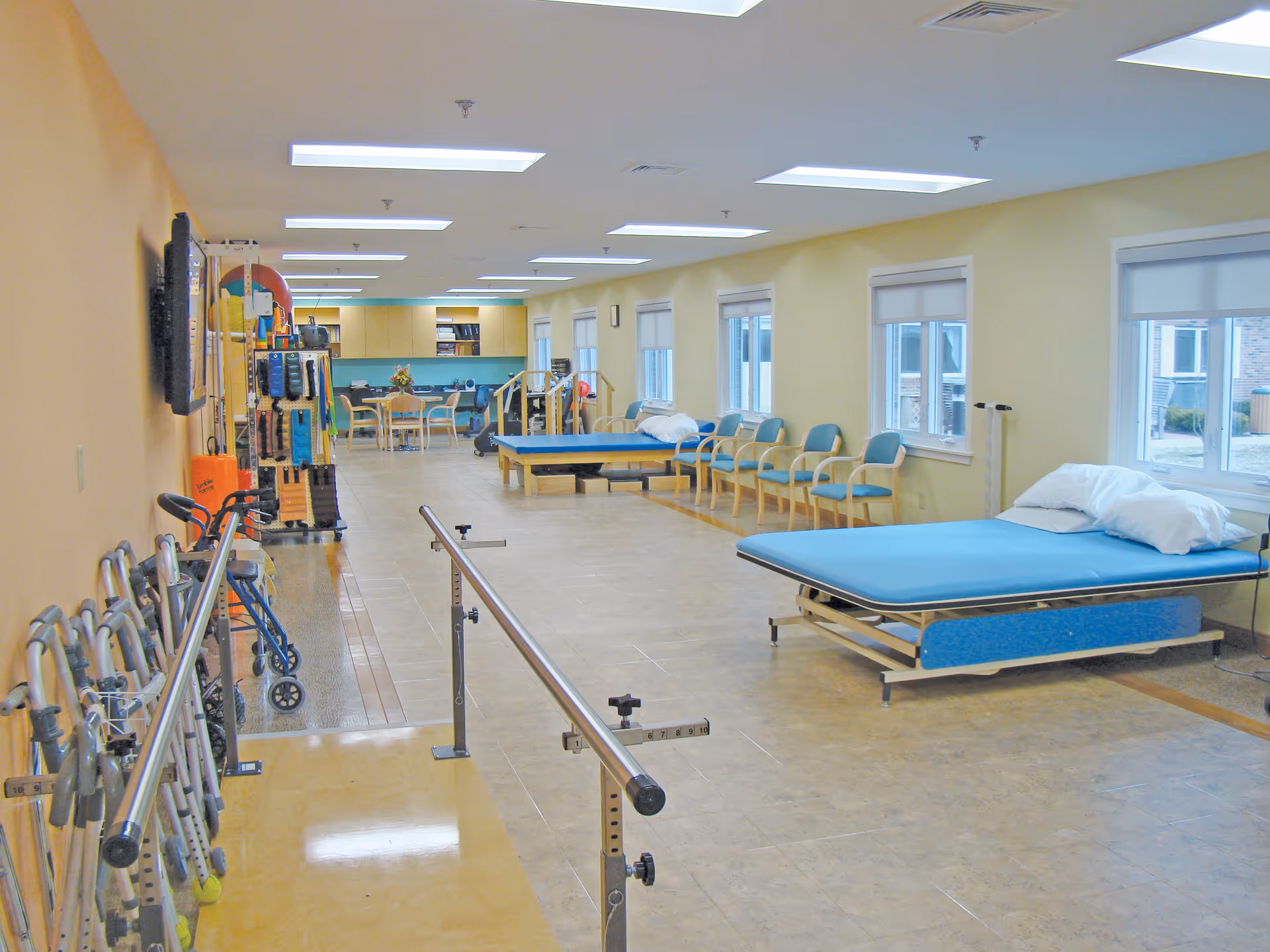 Spacious rehabilitation therapy room with parallel bars, therapy beds, chairs, walkers, and exercise equipment.