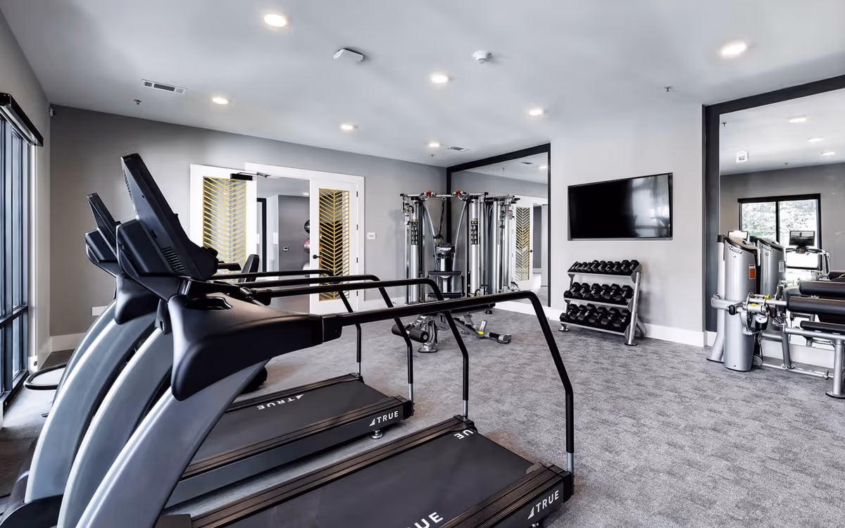 Bright indoor fitness room with treadmills in the foreground, weight rack, strength machines, and a wall-mounted TV.