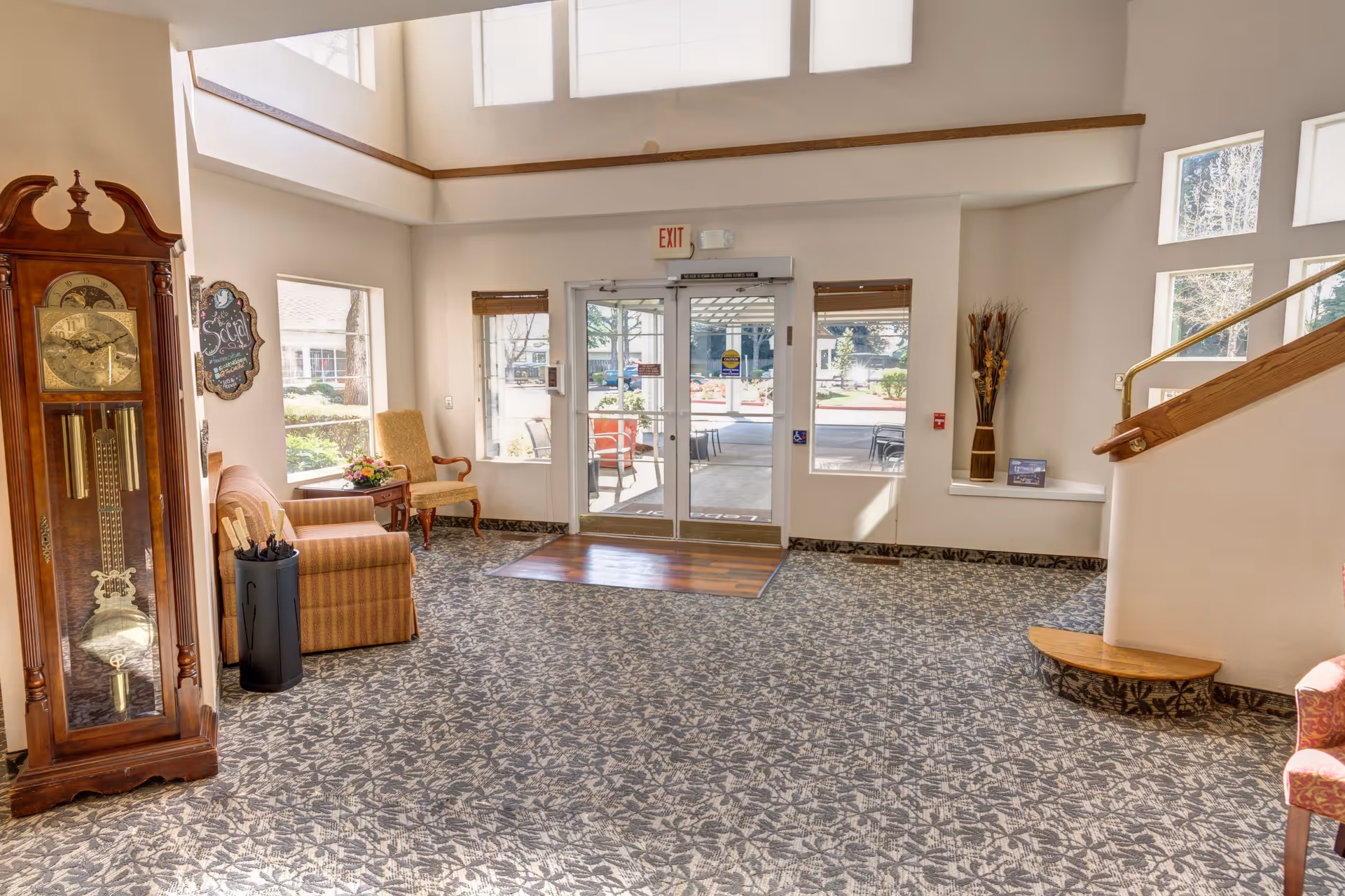 Bright and spacious senior living facility lobby with patterned carpet, a wooden grandfather clock, comfortable armchairs, a small table with flowers, large windows, and glass double doors leading outside. A staircase with a wooden handrail is visible on the right side.