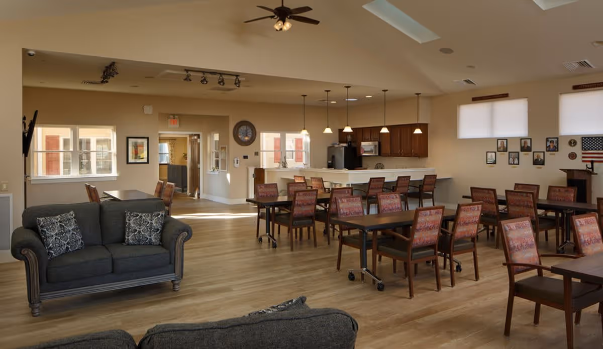 A spacious common area in a senior living facility featuring multiple tables and chairs arranged for dining or activities, a kitchen area with hanging pendant lights and wooden cabinets, and two gray sofas with patterned cushions. The room has large windows allowing natural light, light wood flooring, and framed pictures on the walls.