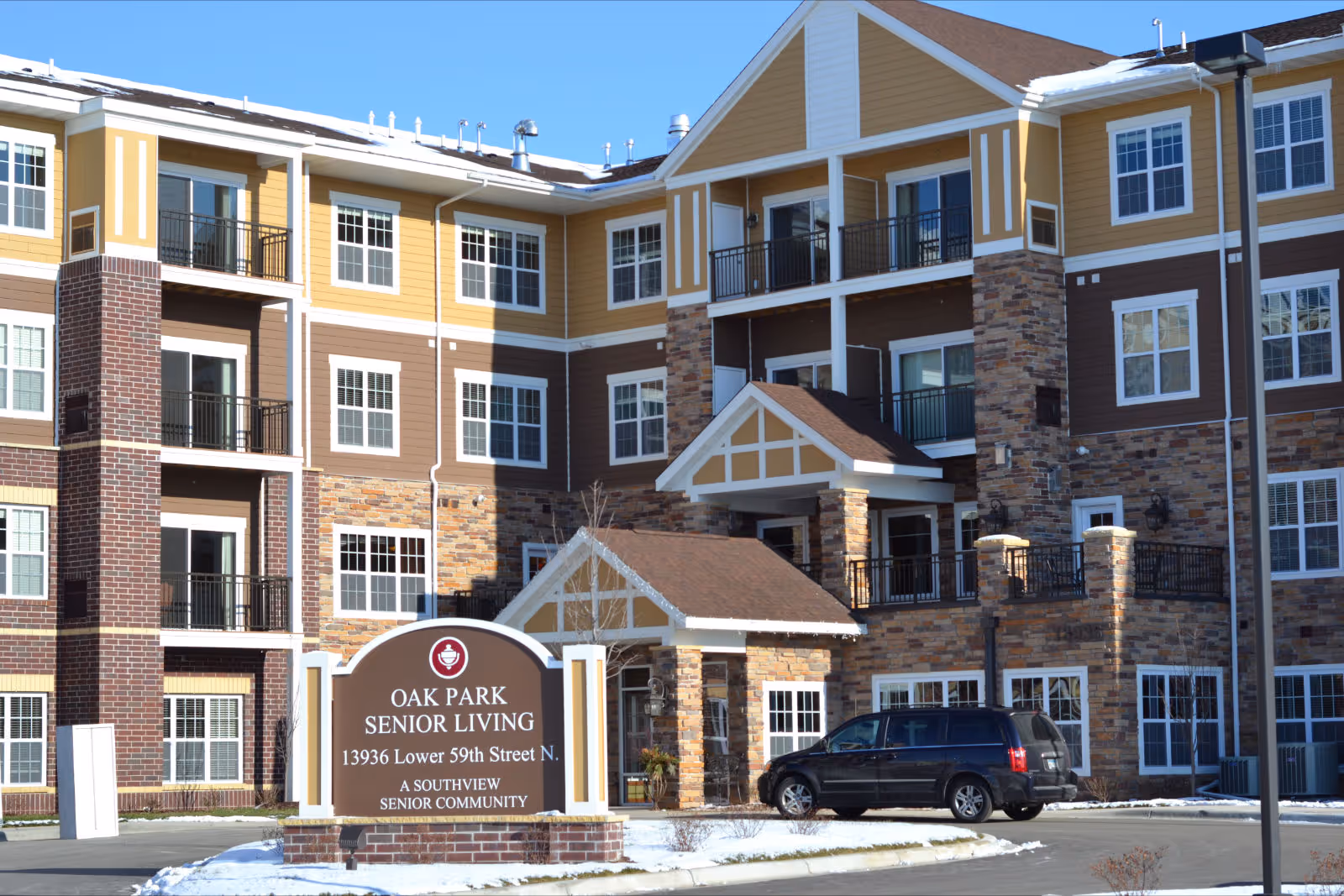 Exterior view of Oak Park Senior Living, a multi-story senior living community building with a mix of brick, stone, and siding facade. The building has multiple balconies and large windows. A sign in front displays the name, address, and description of the community. A black vehicle is parked near the entrance, and there is a light covering of snow on the ground.