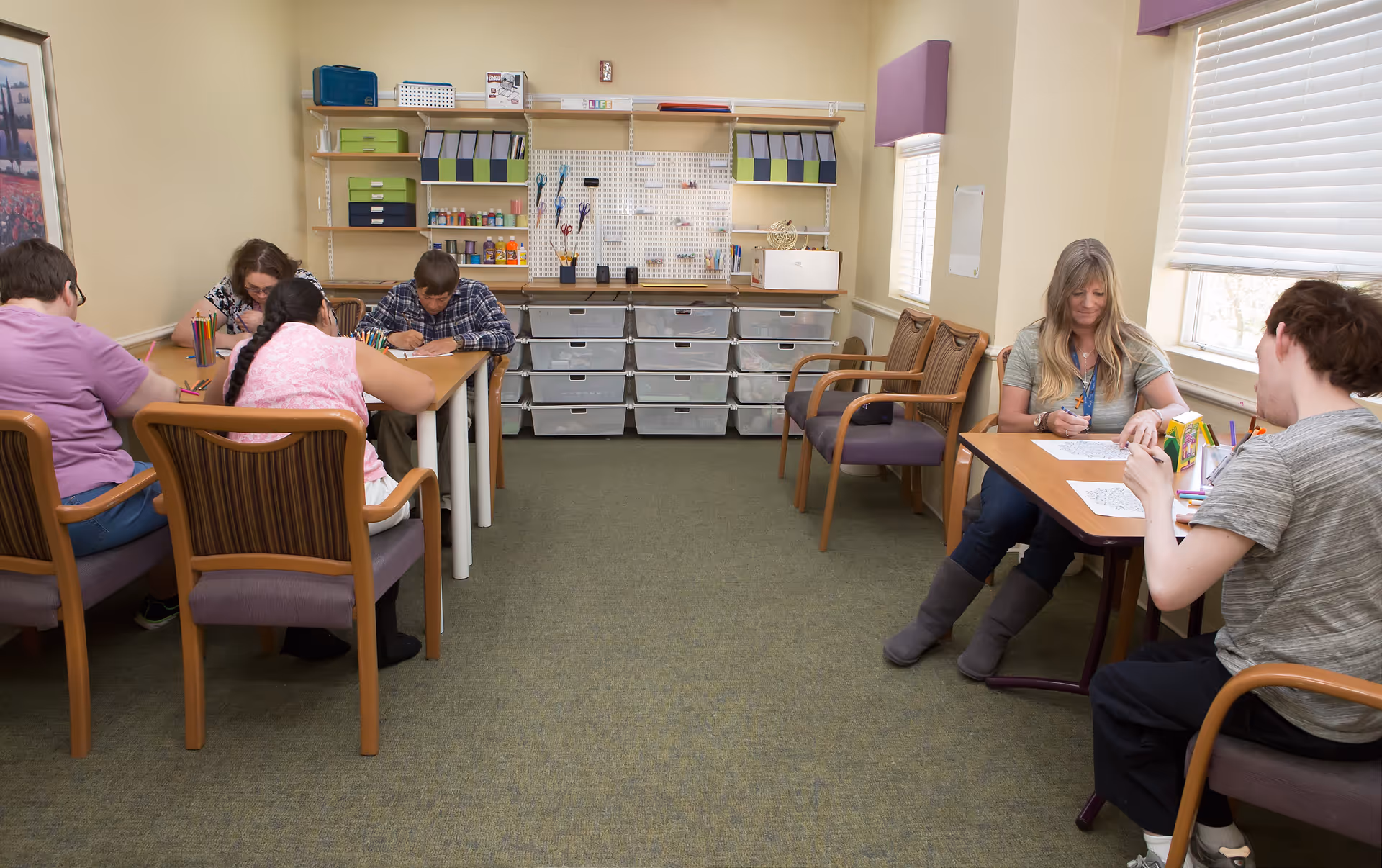 A group of people sitting at two separate tables in a well-lit room, engaged in drawing or coloring activities. The room has beige walls, carpeted floor, shelves with organized supplies, and windows with blinds letting in natural light.