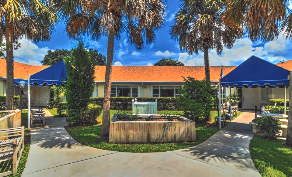 Outdoor courtyard area with palm trees, benches, and a raised garden bed in front of a single-story building with a red roof and blue awnings over the entrances.