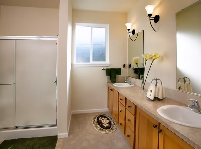 A clean and well-lit bathroom with a double sink vanity featuring wooden cabinets and a beige countertop. Above the sinks are two wall-mounted light fixtures and a large mirror. A vase with yellow flowers and neatly folded towels are placed on the countertop. To the left, there is a glass-enclosed shower with frosted sliding doors. A small window lets in natural light, and green towels and rugs add a touch of color to the neutral-toned room.