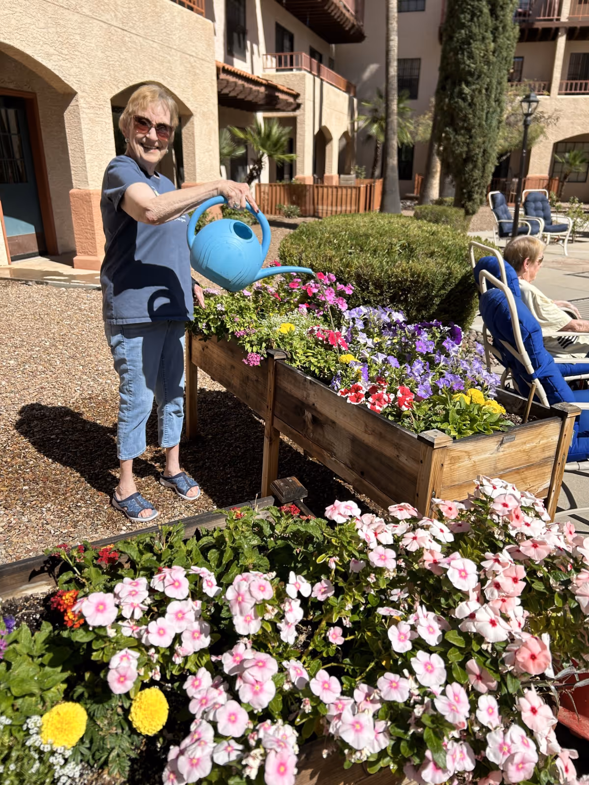 An elderly woman wearing sunglasses and casual clothes waters colorful flowers in a raised wooden garden bed outdoors at a senior living facility. Another elderly person is seated in a blue cushioned chair nearby. The background shows a building with balconies, palm trees, and other outdoor seating.