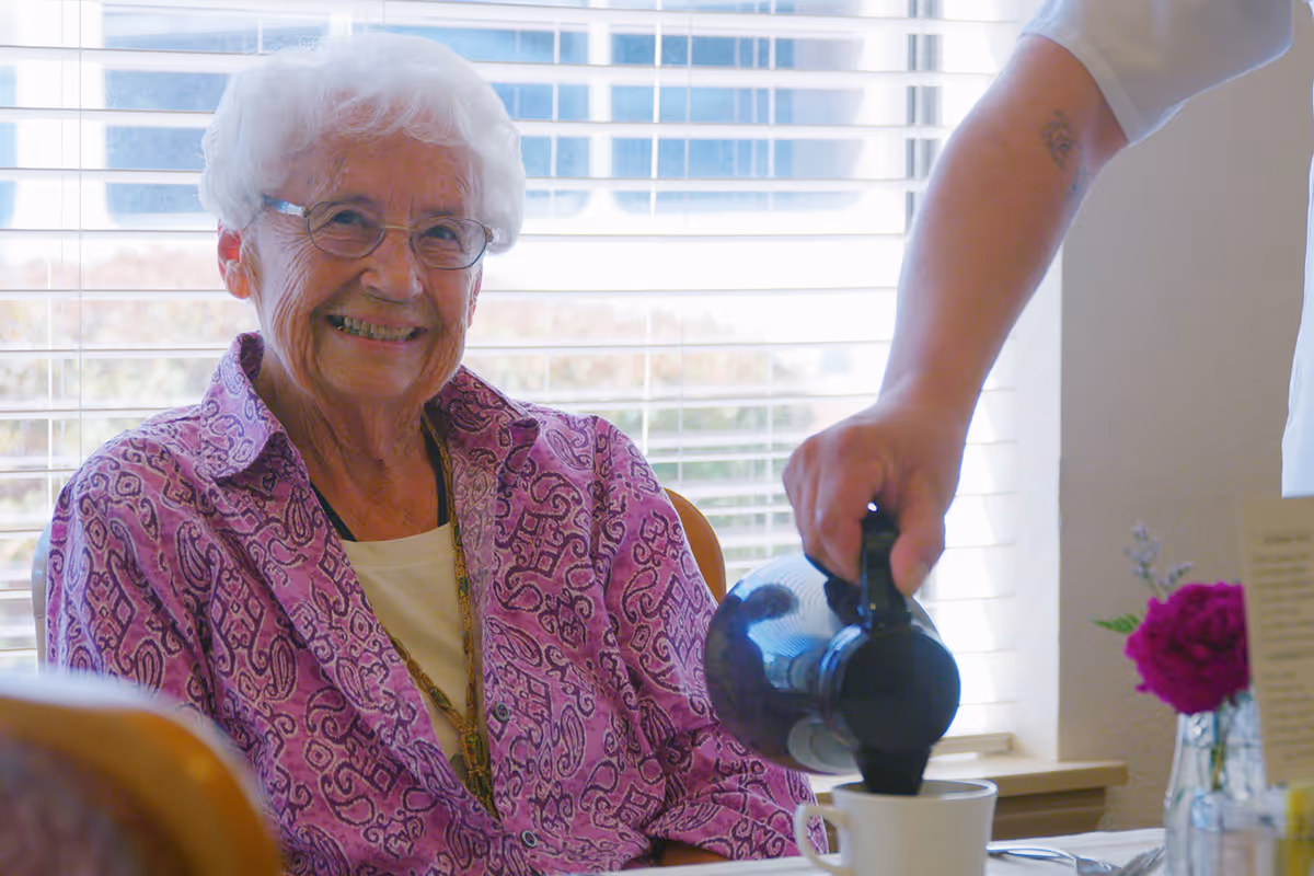 An older woman sitting at a table smiles as someone pours coffee into her cup.