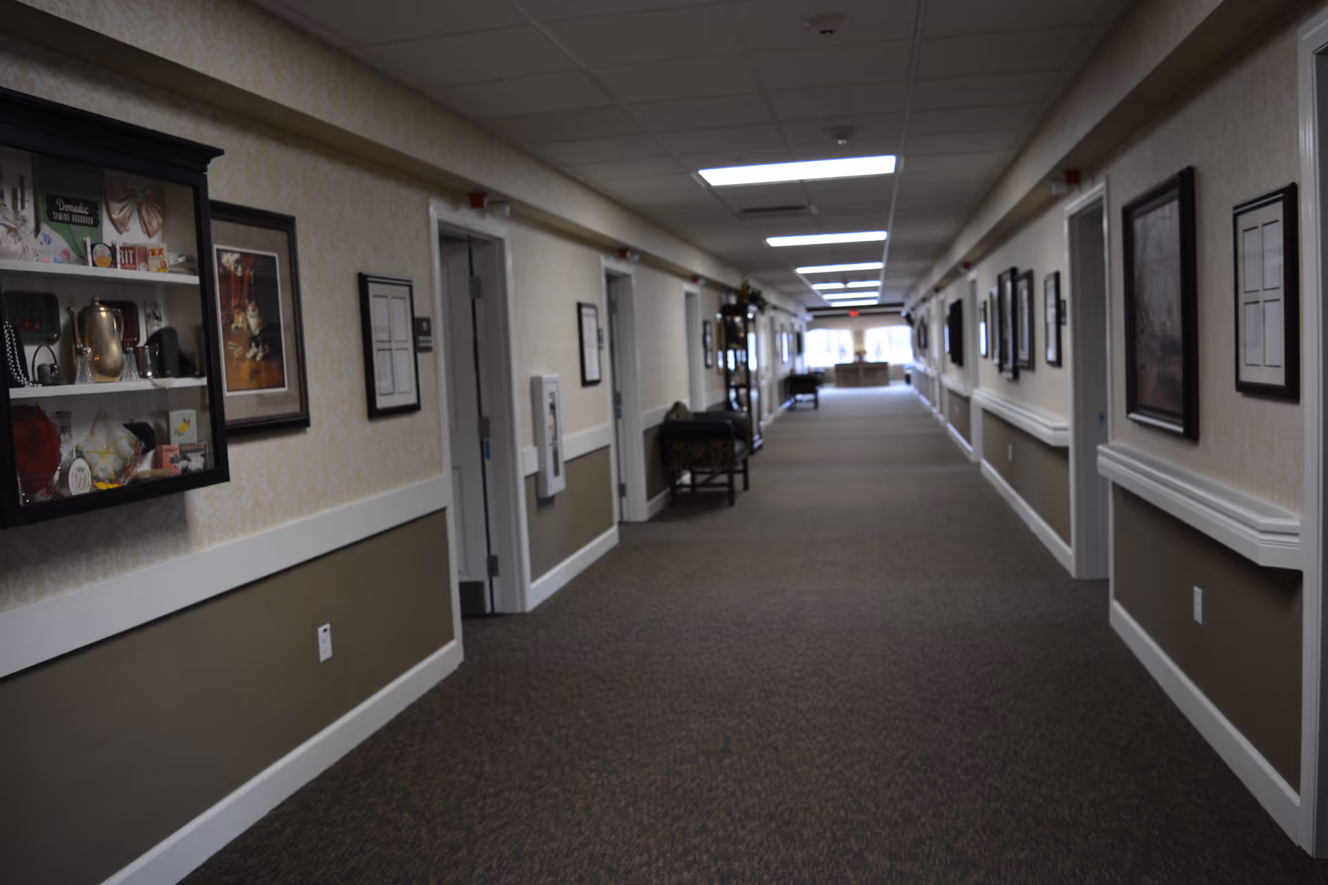A long, carpeted hallway in a senior living facility with beige and brown walls, decorated with framed pictures and display cases. Several doors line the hallway, and there are chairs and small tables placed along the walls. The ceiling has recessed lighting panels.