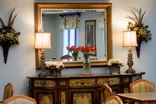 An elegant interior room featuring a large ornate mirror above a decorative wooden sideboard. The sideboard is adorned with a vase of red flowers in the center and smaller floral arrangements on either side. Two matching table lamps with beige shades are positioned symmetrically on the sideboard. The room includes upholstered chairs and a table, with soft lighting creating a warm and inviting atmosphere.