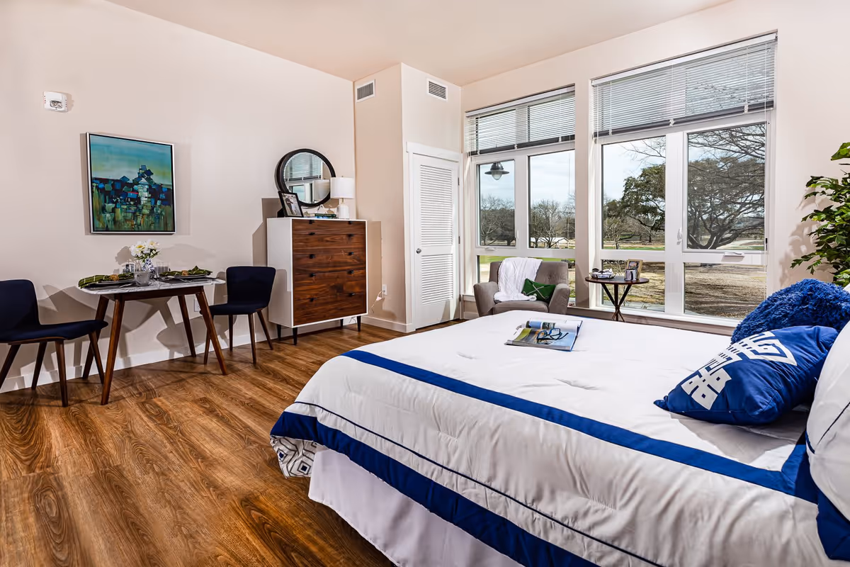 Bright furnished bedroom with a bed, small dining table, dresser, armchair and large windows overlooking trees.